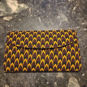 Puede incluir: Un bolso de mano rectangular con solapa. La tela presenta un patrón repetitivo de formas amarillas y granates sobre un fondo negro. El bolso está diseñado para llevar objetos pequeños.