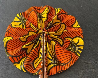 May include: A hand fan with a vibrant, patterned design in yellow, orange, and black. The fan's fabric is pleated and attached to a brown wooden handle with a leather strap. The fan is open, displaying its full circular shape.