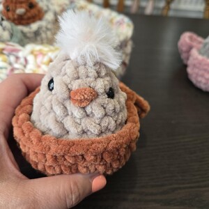 May include: A crocheted chick with a white fluffy pom-pom on its head, sitting in a brown crocheted nest.