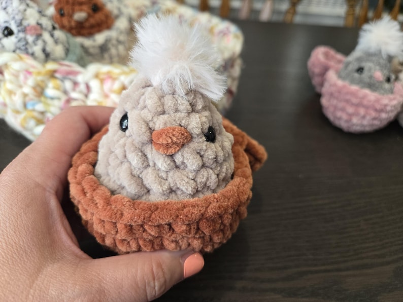 May include: A crocheted chick with a white fluffy pom-pom on its head, sitting in a brown crocheted nest.