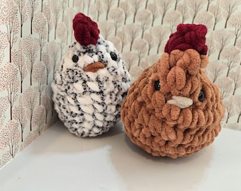 May include: Two plush crochet chickens made from soft yarn. One chicken is white with black speckles, and the other is solid brown. Both feature a dark red textured comb on top, small black bead eyes, and a light-colored beak. The figures are rounded.
