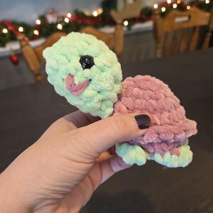 May include: A crocheted turtle toy with a green head and pink shell. The turtle has black eyes and a pink mouth.