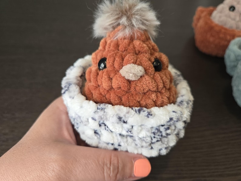 May include: A small, brown crocheted chick with black eyes and a fluffy gray pom-pom on its head. The chick is sitting in a white and gray crocheted nest.