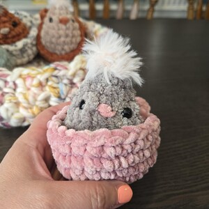 May include: A gray crocheted chick with a white fluffy crest sits in a pink crocheted nest.