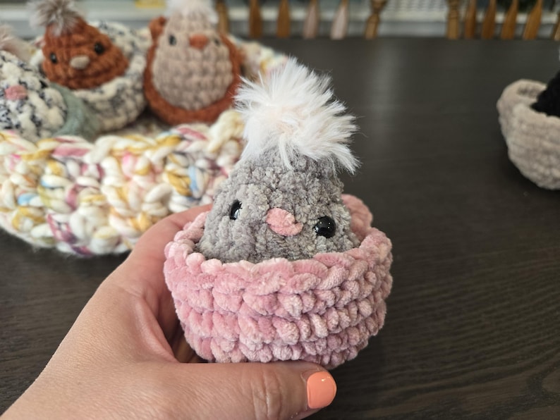 May include: A gray crocheted chick with a white fluffy crest sits in a pink crocheted nest.