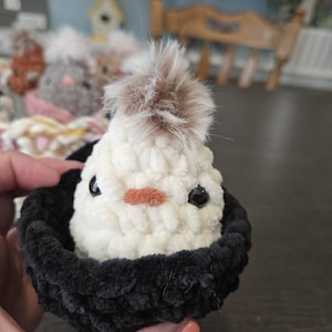 May include: A white crocheted chick with brown fur on top of its head, sitting in a black crocheted nest.