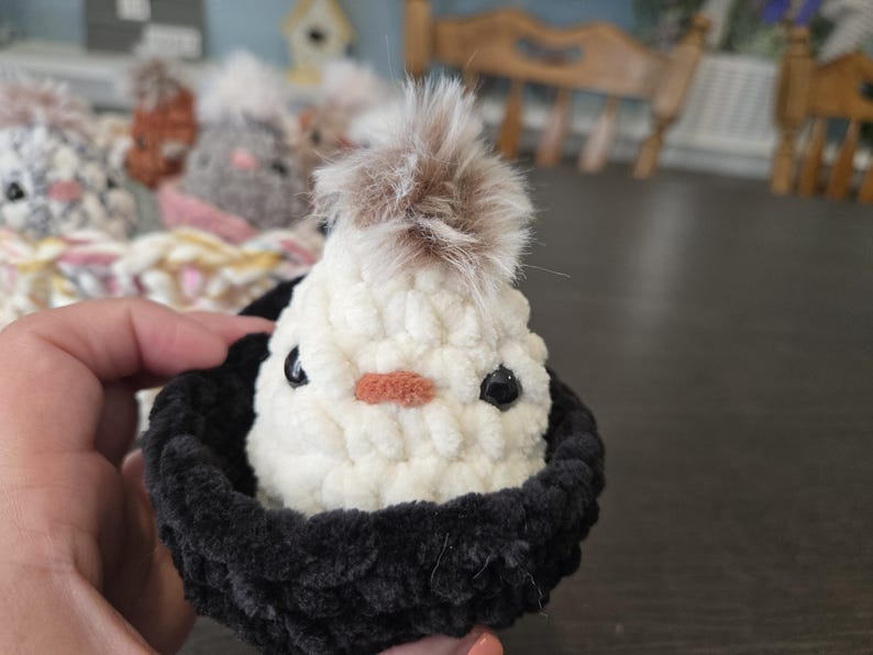 May include: A white crocheted chick with brown fur on top of its head, sitting in a black crocheted nest.