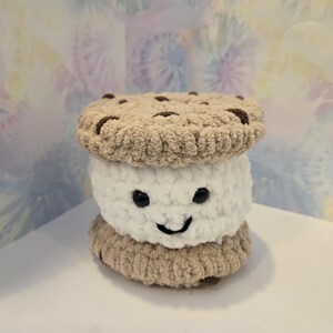 May include: A handmade, plush cookie sandwich with a smiling face. The top and bottom layers are tan, resembling cookies with brown chocolate chips. The middle layer is white, with black eyes and a stitched smile.