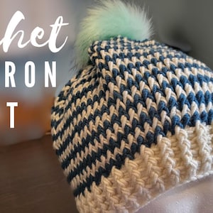 Puede incluir: Un gorro de punto chevron con un patrón de chevron blanco y azul. El gorro tiene un pompón de color azul claro y esponjoso en la parte superior.