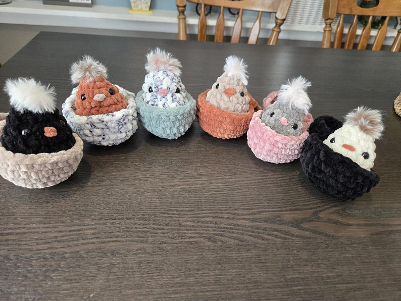 May include: Six crocheted chicks in nests, each with a different colored nest and chick. The chicks are all in various poses, with some looking up and some looking down. The nests are all made of a soft, fuzzy yarn.
