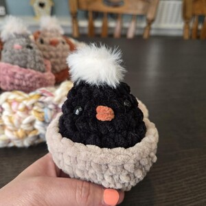 May include: A black crocheted chick with a white fluffy pom-pom on its head. The chick is sitting in a beige crocheted nest.