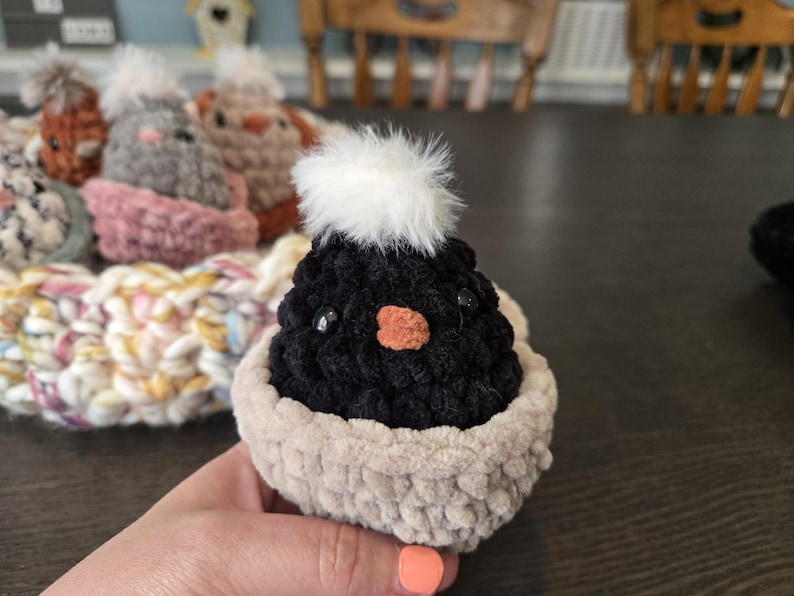 May include: A black crocheted chick with a white fluffy pom-pom on its head. The chick is sitting in a beige crocheted nest.