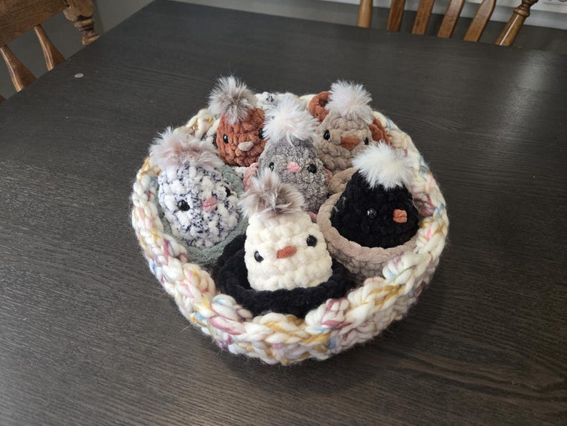 May include: A crocheted basket filled with seven small crocheted chicks. The chicks are in various colors, including white, brown, gray, and black. The basket is made of a variety of colors, including pink, white, and yellow.