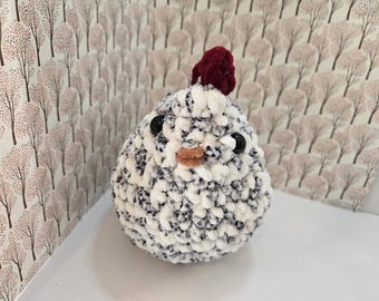 May include: A white and gray crocheted chick toy with black eyes and a red felt comb. The chick is sitting on a white surface with a patterned background.