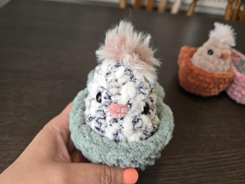 May include: A small, crocheted bird with a pink and white fuzzy head, sitting in a green crocheted nest.