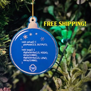 May include: A blue Christmas bauble with a white snowflake design and code printed on it. The bauble is hanging from a gold string on a green Christmas tree. The text on the bauble reads: "void setup() { pinMode(13, OUTPUT); } void loop() { digitalWrite(13, HIGH); delay(1000); digitalWrite(13, LOW); delay(1000); "