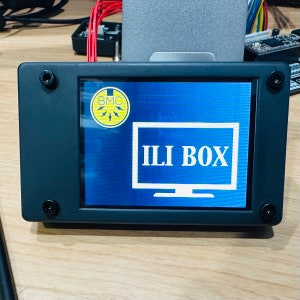 Op de afbeelding: Een zwart omlijst scherm met een blauwe achtergrond waarop de tekst "ILI BOX" in wit wordt weergegeven. Een gele sticker met de tekst "BMC" in zwart bevindt zich in de linkerbovenhoek van het scherm.