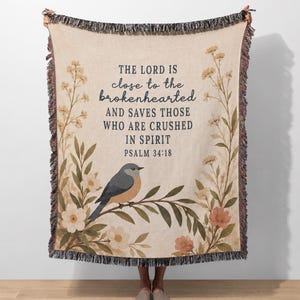 May include: A woven throw blanket in a light beige color featuring a blue and orange bird perched on a branch. The blanket displays the text THE LORD IS close to the brokenhearted AND SAVES THOSE WHO ARE CRUSHED IN SPIRIT PSALM 34:18.