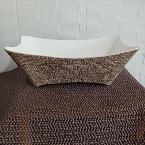 May include: A rectangular serving bowl with a white interior and a textured brown exterior. The bowl has a unique shape with flared sides. The exterior has a woven pattern. The bowl is sitting on a brown surface.