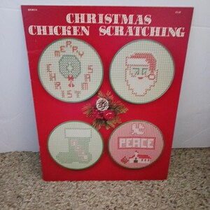 May include: A red instructional booklet titled CHRISTMAS CHICKEN SCRATCHING featuring four circular cross-stitch patterns: a wreath with MERRY CHRISTMAS, a Santa face, a stocking, and a house with the word PEACE, plus a small pinecone decoration.