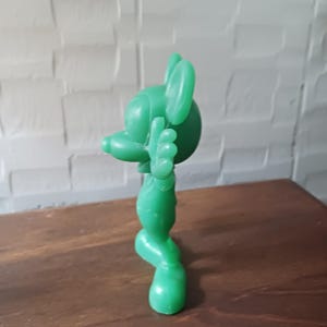 May include: A solid green figurine of a cartoon mouse with large ears, a long nose, and outstretched arms. The figure is standing on a wooden surface, with a white brick wall in the background.
