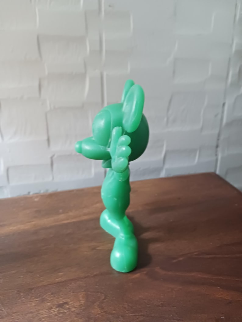May include: A solid green figurine of a cartoon mouse with large ears, a long nose, and outstretched arms. The figure is standing on a wooden surface, with a white brick wall in the background.