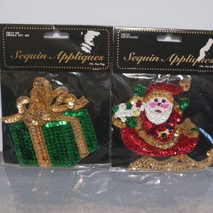 May include: Two sequin appliqués in clear plastic packaging. The left appliqué is a green gift box with a gold bow. The right appliqué features Santa Claus in a red suit riding a gold sleigh. Both packages are labeled Sequin Appliques.