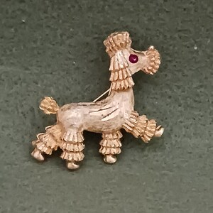 May include: A gold-toned poodle brooch with a red rhinestone eye. The poodle is standing on all four legs with its tail curled up.