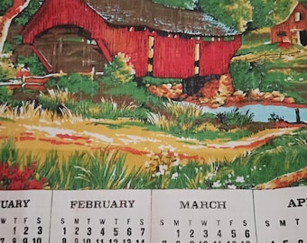May include: A fabric with a scenic print featuring a red covered bridge, trees, and a stream. The bottom of the fabric has a calendar with the months of February and March visible.