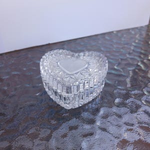 May include: A clear, heart-shaped trinket box with a frosted heart design on the lid. The box is made of cut crystal, showcasing a geometric pattern. The box is sitting on a textured surface.