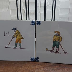 May include: Two white ceramic tiles with hand-painted illustrations of golfers. Each figure is dressed in yellow and green attire, playing golf. The tiles have blue floral corner accents.