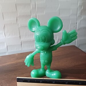 May include: A green Mickey Mouse figurine with outstretched arms and a friendly expression. The figure is standing on a wooden surface, with a ruler placed nearby. The figurine is approximately 4 inches tall.