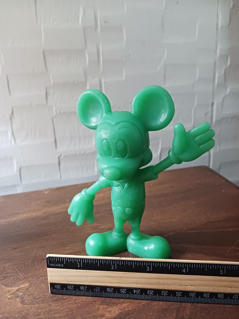 May include: A green Mickey Mouse figurine with outstretched arms and a friendly expression. The figure is standing on a wooden surface, with a ruler placed nearby. The figurine is approximately 4 inches tall.