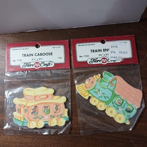 May include: Two decorative wooden train cutouts, a caboose and an engine, each in a plastic bag. The caboose is peach, yellow, and blue, while the engine is green, peach, and yellow. Each is 3.75" x 2.25" and labeled "Decorative Use Only."