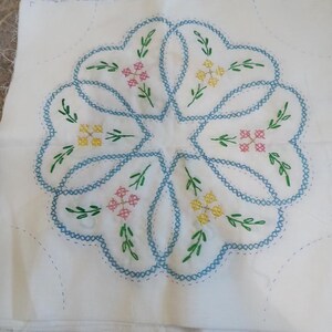 May include: A square white fabric piece featuring a central embroidered floral motif. The design includes blue cross-stitched scalloped outlines, green leaves, and small yellow and pink flower clusters. A wooden ruler is visible at the bottom edge.