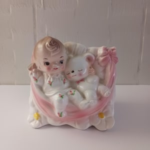 May include: A vintage ceramic planter featuring a sleeping baby and a teddy bear. The planter is primarily white with pink accents, including a ribbon and a cradle-like design. The baby and bear are white with delicate facial features and details.