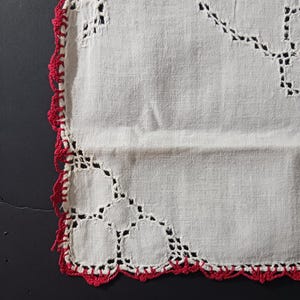 May include: White linen doily with a red crocheted border. The doily has a delicate lace pattern.