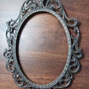 May include: An oval-shaped metal picture frame featuring an intricate, ornate border with scrolling filigree and leaf-like patterns. The frame has a dark, weathered finish with subtle gold-toned highlights along the raised decorative edges.