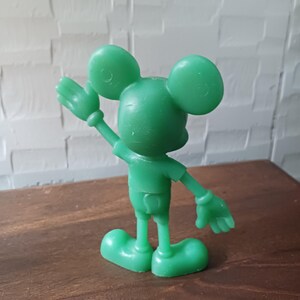 May include: A green plastic figurine of a cartoon mouse with large round ears, wearing shorts and a short-sleeved shirt. The mouse is waving with one hand raised. The figurine is standing on a brown surface.