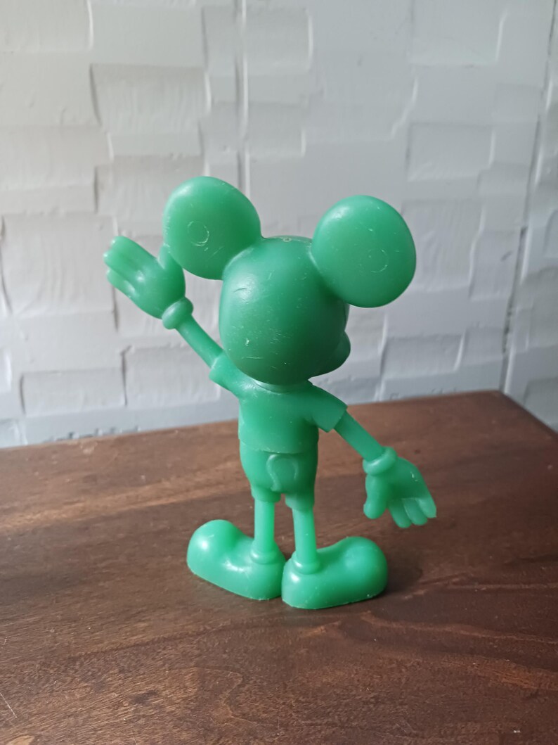 May include: A green plastic figurine of a cartoon mouse with large round ears, wearing shorts and a short-sleeved shirt. The mouse is waving with one hand raised. The figurine is standing on a brown surface.