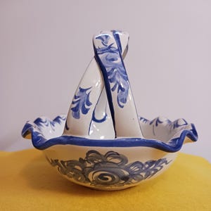 May include: A white ceramic basket with a blue and white floral design. The basket has a twisted handle and a scalloped edge.