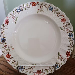May include: White ceramic plate with a scalloped edge, decorated with a floral pattern in red, blue, and green. The plate has a textured surface and is displayed on a metal plate stand. A decorative piece for the dining table.