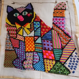 May include: A needlepoint tapestry featuring a black cat composed of various colorful geometric and floral patterns. The cat has green eyes, a pink nose, and a yellow bow tie, set against a cream-colored mesh canvas background with visible edges.