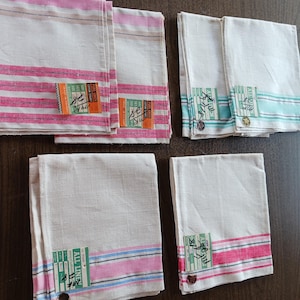 May include: A collection of white linen handkerchiefs with colorful striped borders in pink, blue, and green. Each handkerchief has a rectangular label with Japanese text and a button. The handkerchiefs are arranged on a dark brown surface.