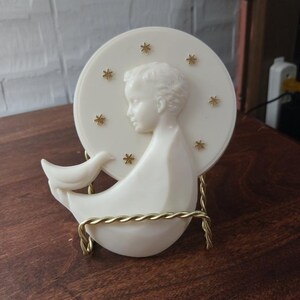 May include: A white ceramic sculpture of a child in profile, holding a dove, set against a circular backdrop with gold star accents. The sculpture is supported by a gold wire stand. The piece is displayed on a wooden surface.