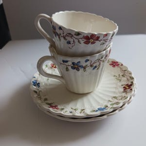 May include: Two cream-colored ceramic teacups with scalloped edges and ribbed sides, stacked vertically. The cups and matching saucers feature a delicate floral pattern with red and blue flowers and green vines painted along the rims.