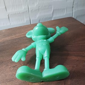 May include: A solid green figurine of a cartoon mouse with large ears, wearing overalls and large shoes. The mouse is posed with one arm raised, and the other arm is at its side. The figurine is on a brown wooden surface.