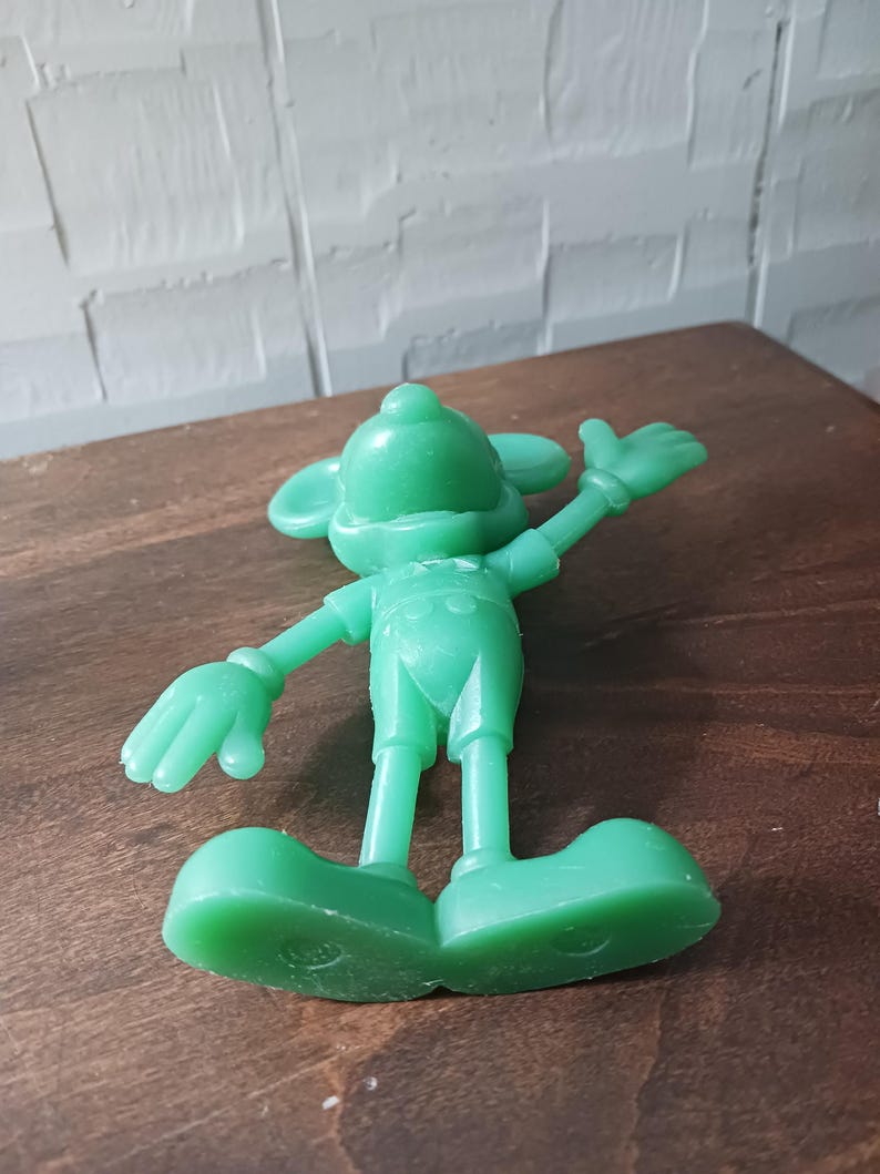 May include: A solid green figurine of a cartoon mouse with large ears, wearing overalls and large shoes. The mouse is posed with one arm raised, and the other arm is at its side. The figurine is on a brown wooden surface.