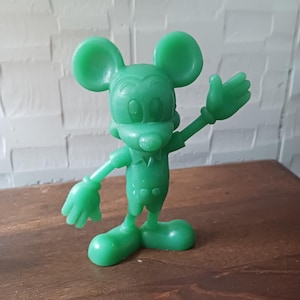 May include: A solid green Mickey Mouse figurine in a waving pose. The figure has large, round ears, a smiling face, and is wearing a short-sleeved shirt and shorts. The figurine is made of a smooth, glossy material.