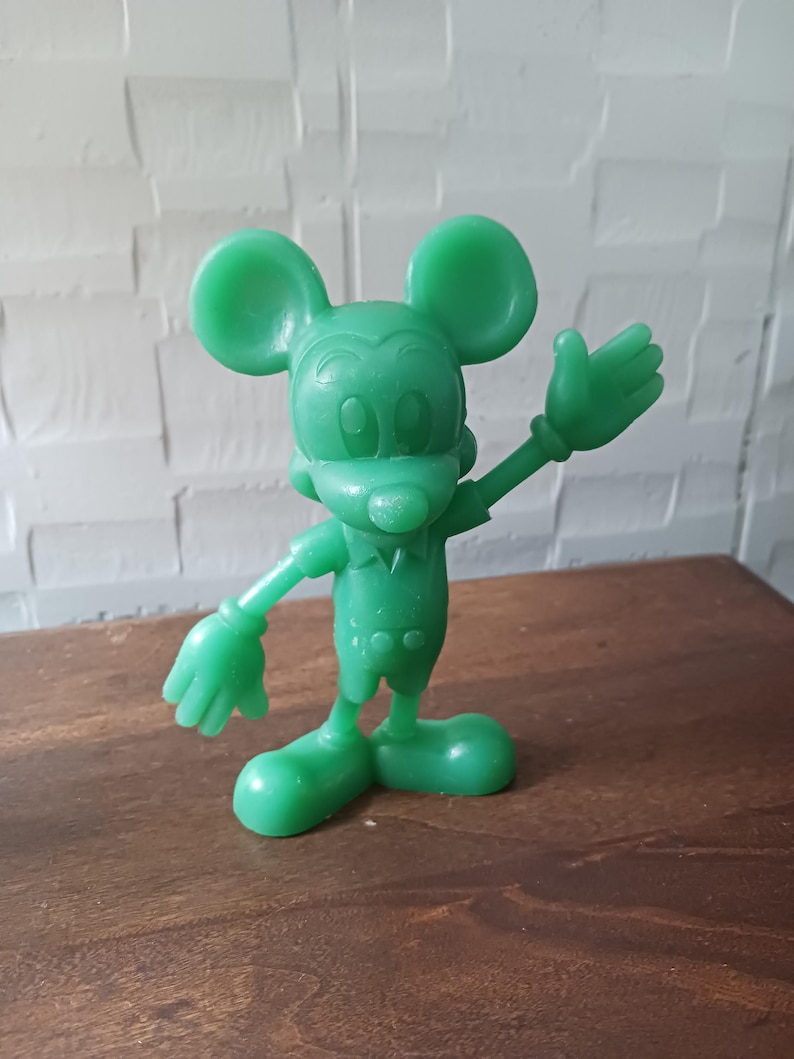 May include: A solid green Mickey Mouse figurine in a waving pose. The figure has large, round ears, a smiling face, and is wearing a short-sleeved shirt and shorts. The figurine is made of a smooth, glossy material.